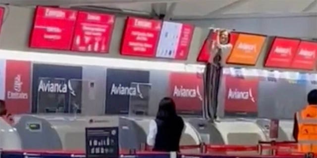 Penumpang lain di bandara dapat melihat orang yang tidak terkendali - berdiri di konter check-in dan memegang layar di atasnya - dari jauh.